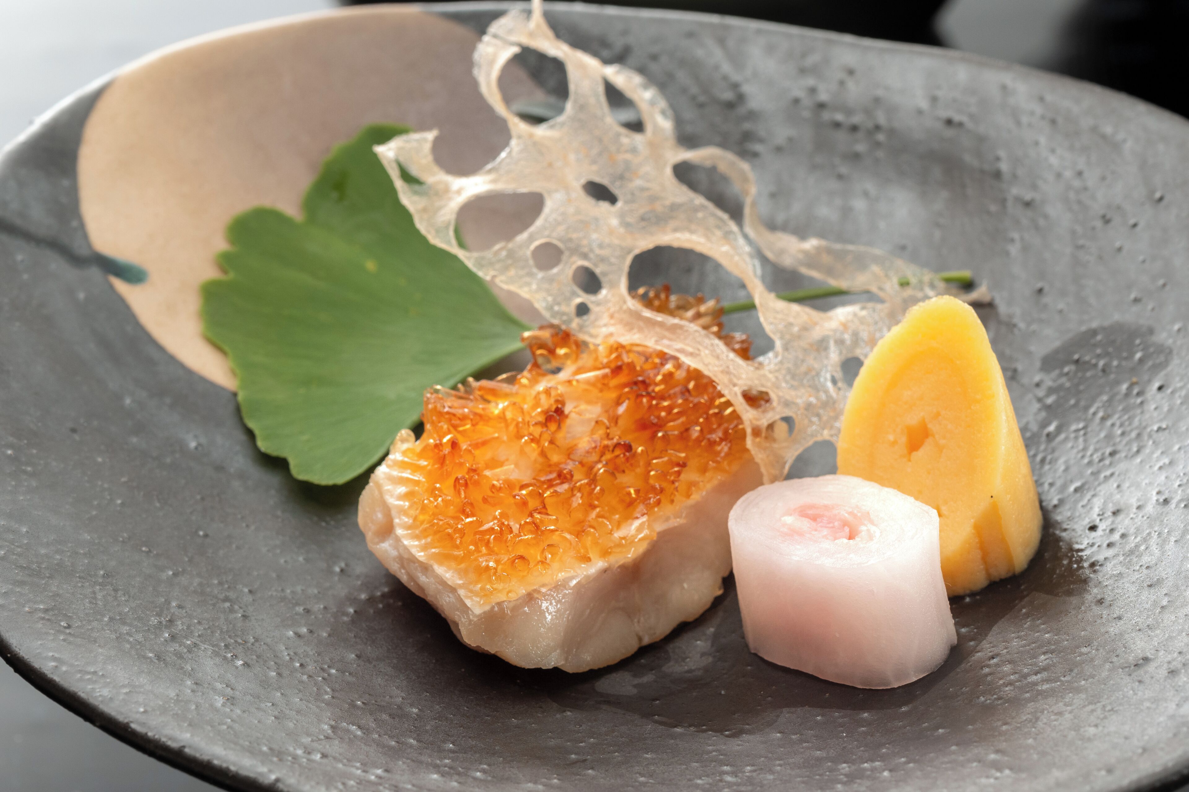 A dish with a piece of fish, a slice of eggplant, and a leaf on a dark plate.