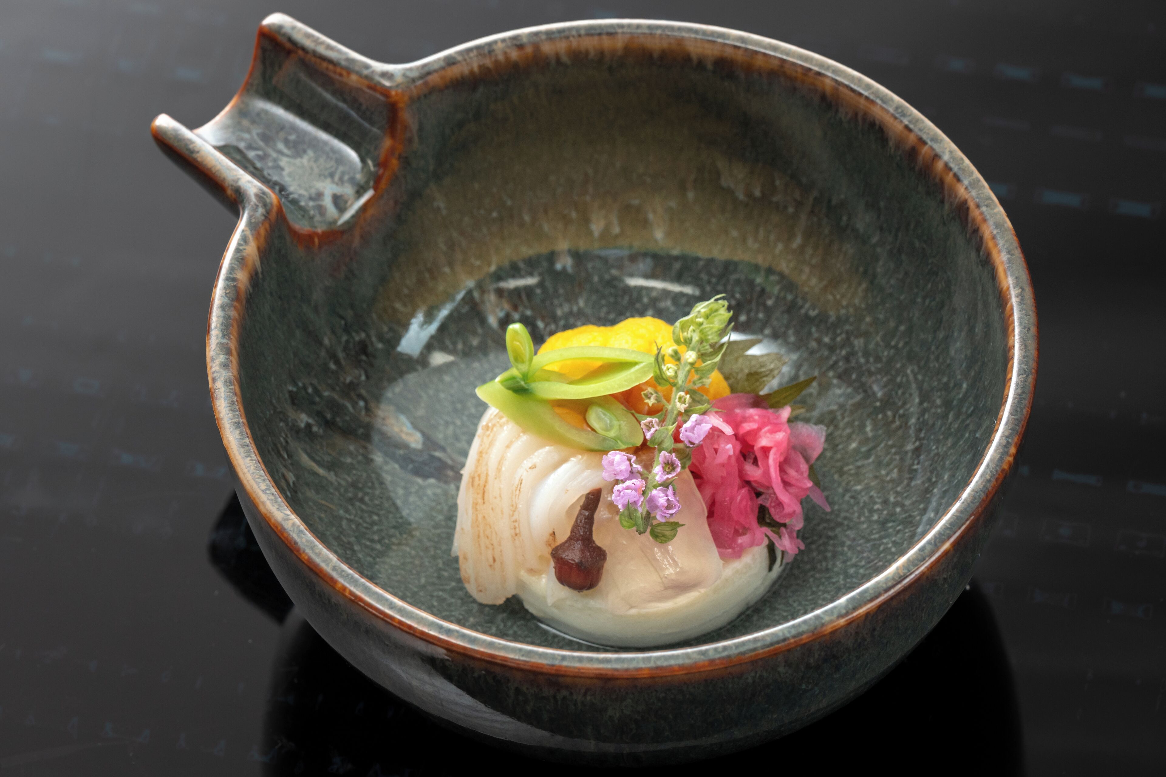 A ceramic bowl with a handle, containing a white food item, green vegetables, and pink flowers.