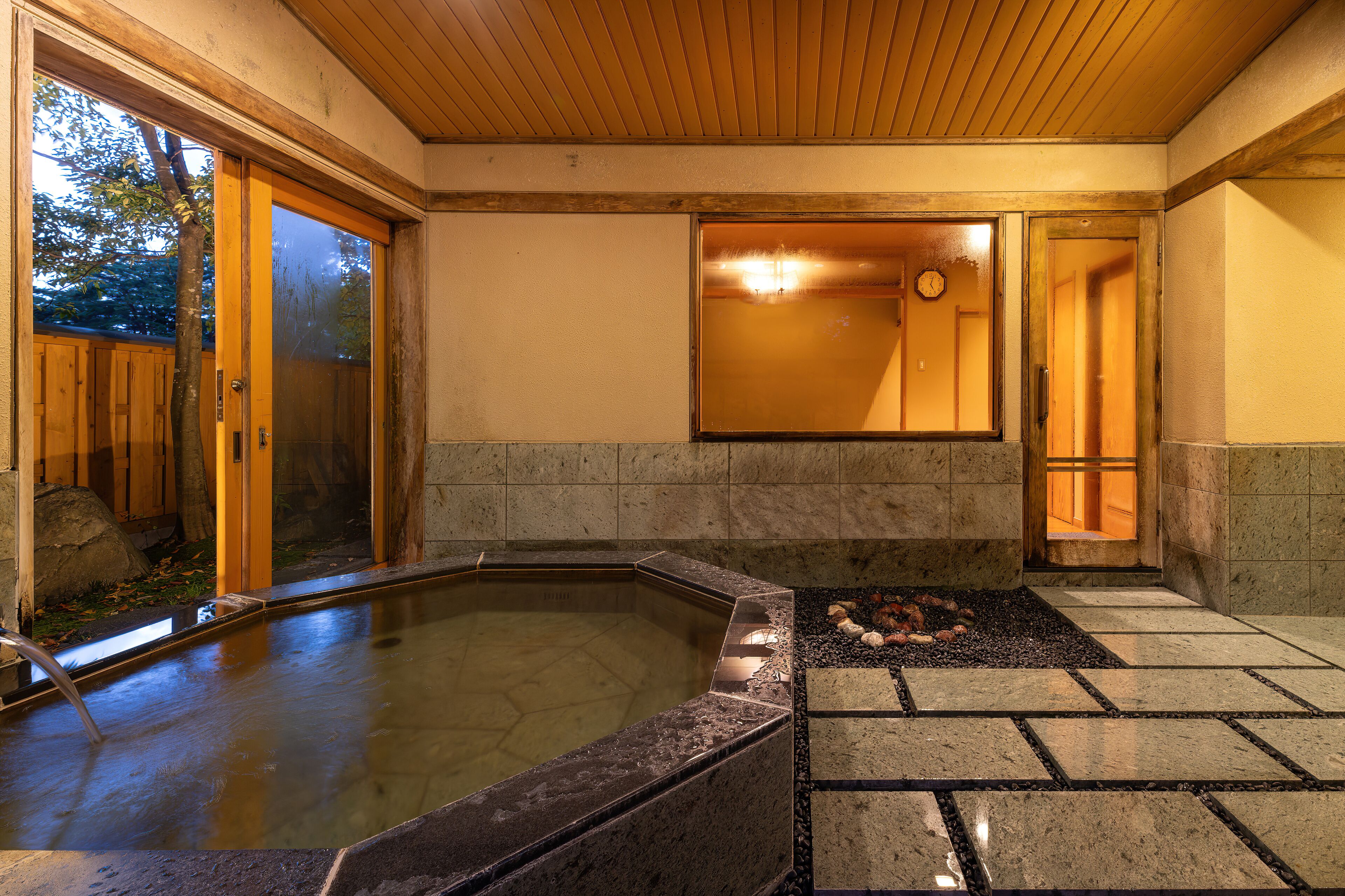 Un onsen japonais traditionnel, avec un intérieur en bois, un sol en pierre et une vue sur l’extérieur grâce à une porte coulissante.