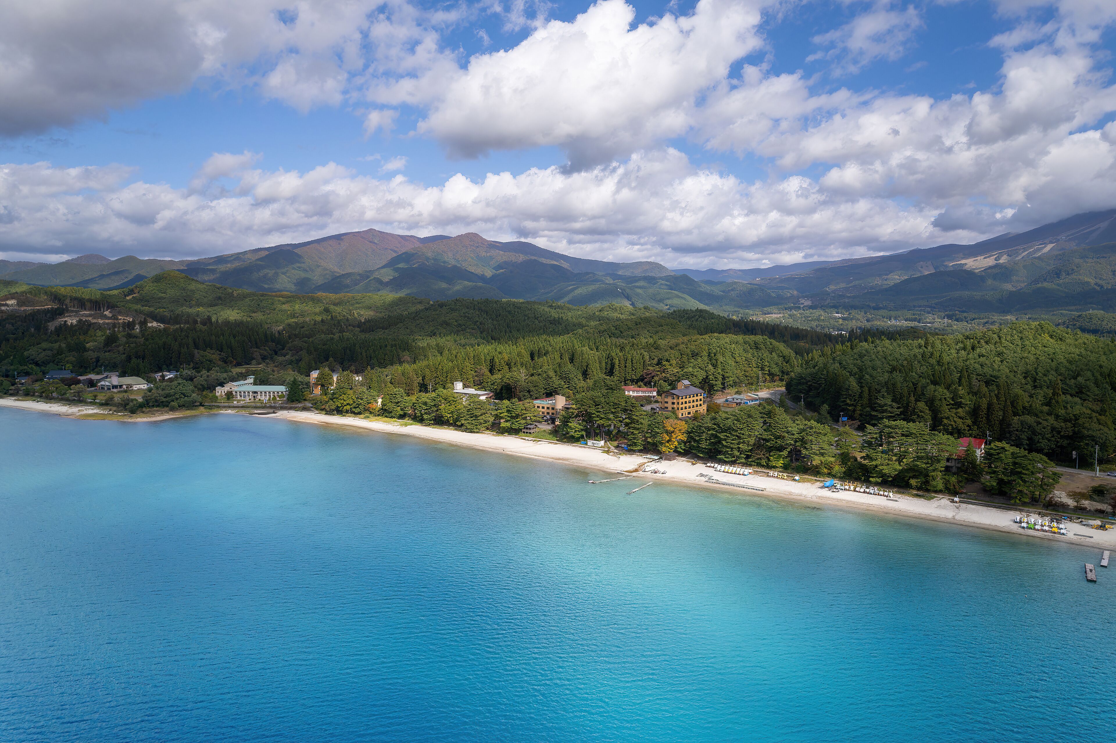 A coastal landscape with a beach, forested hills, and a mountain range in the background.