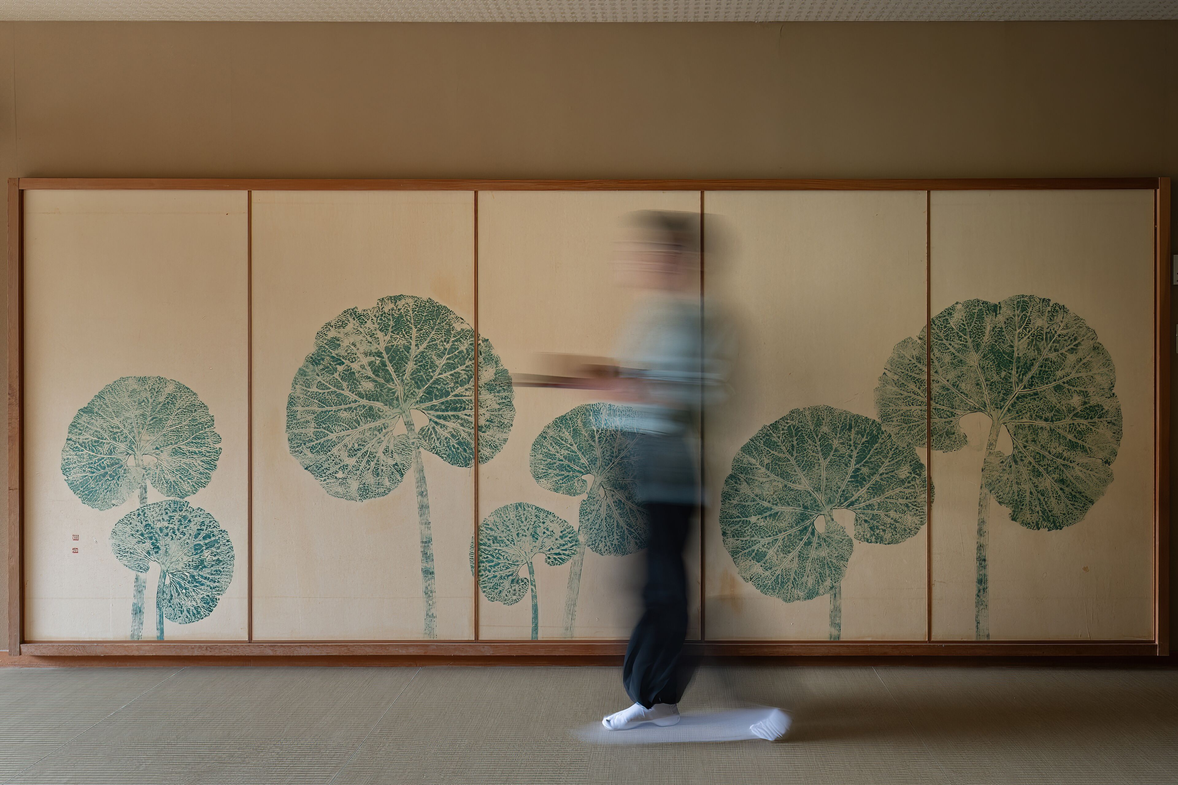Een persoon doet een rekoefening op een mat voor een traditionele Japanse kamer met tatami vloeren en een schuifdeur met bladmotief.