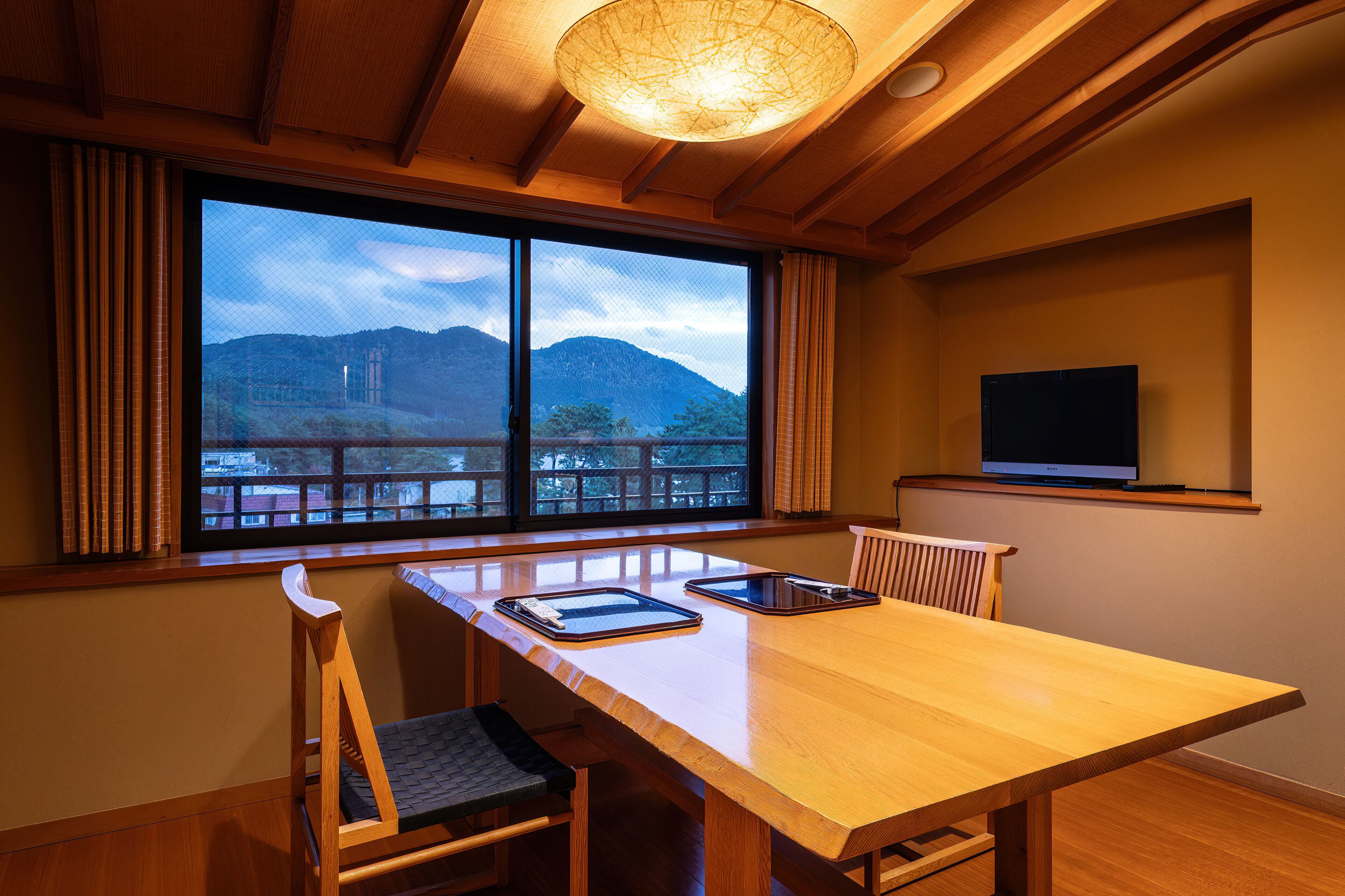 Ein Zimmer mit großem Fenster und Blick auf Berge und See, einem Holztisch mit Stühlen, einem an der Wand montierten Fernseher und einer Deckenleuchte.