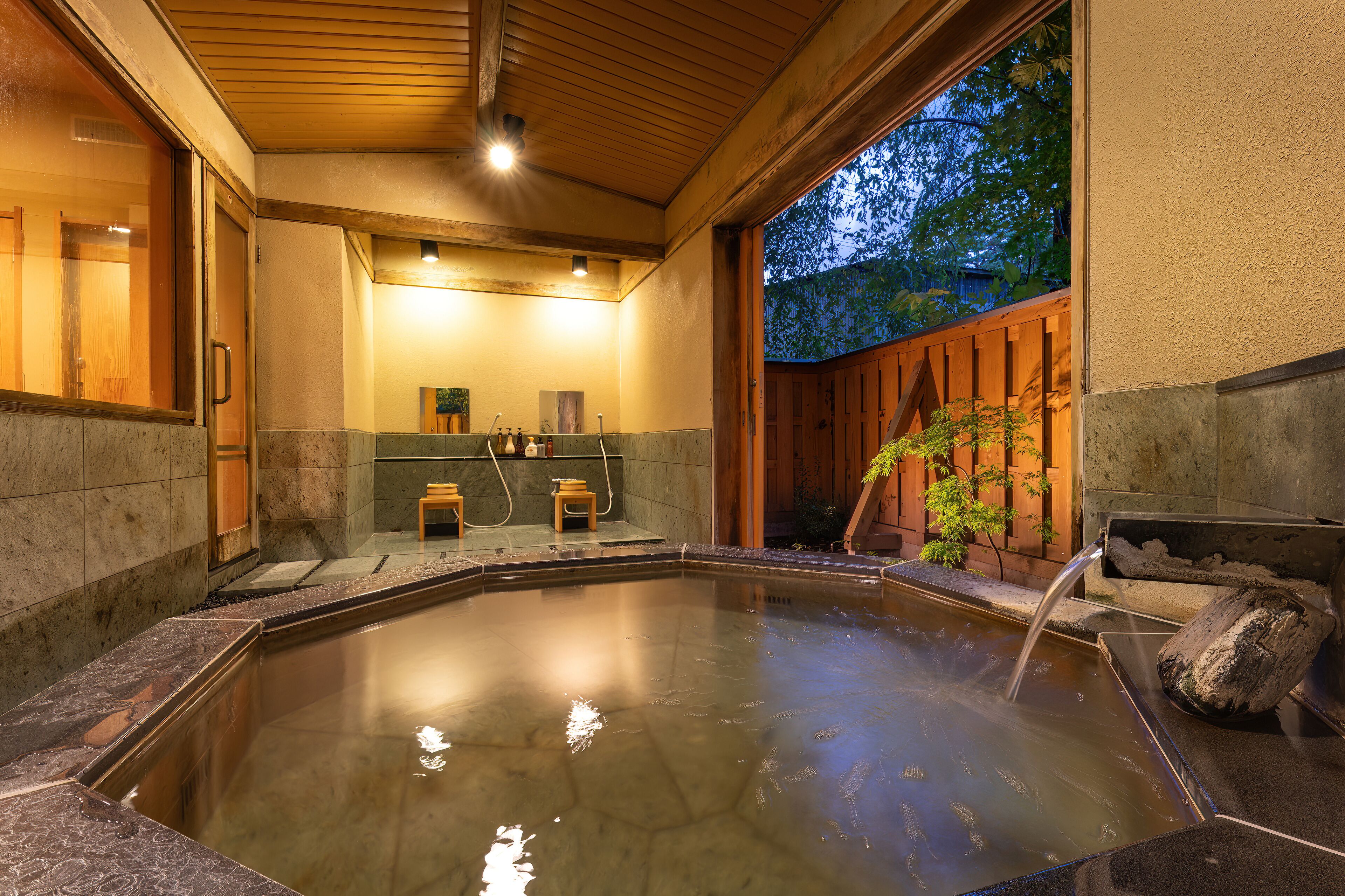 A hot spring bath area with a wooden ceiling, stone walls, and a view of trees outside.
