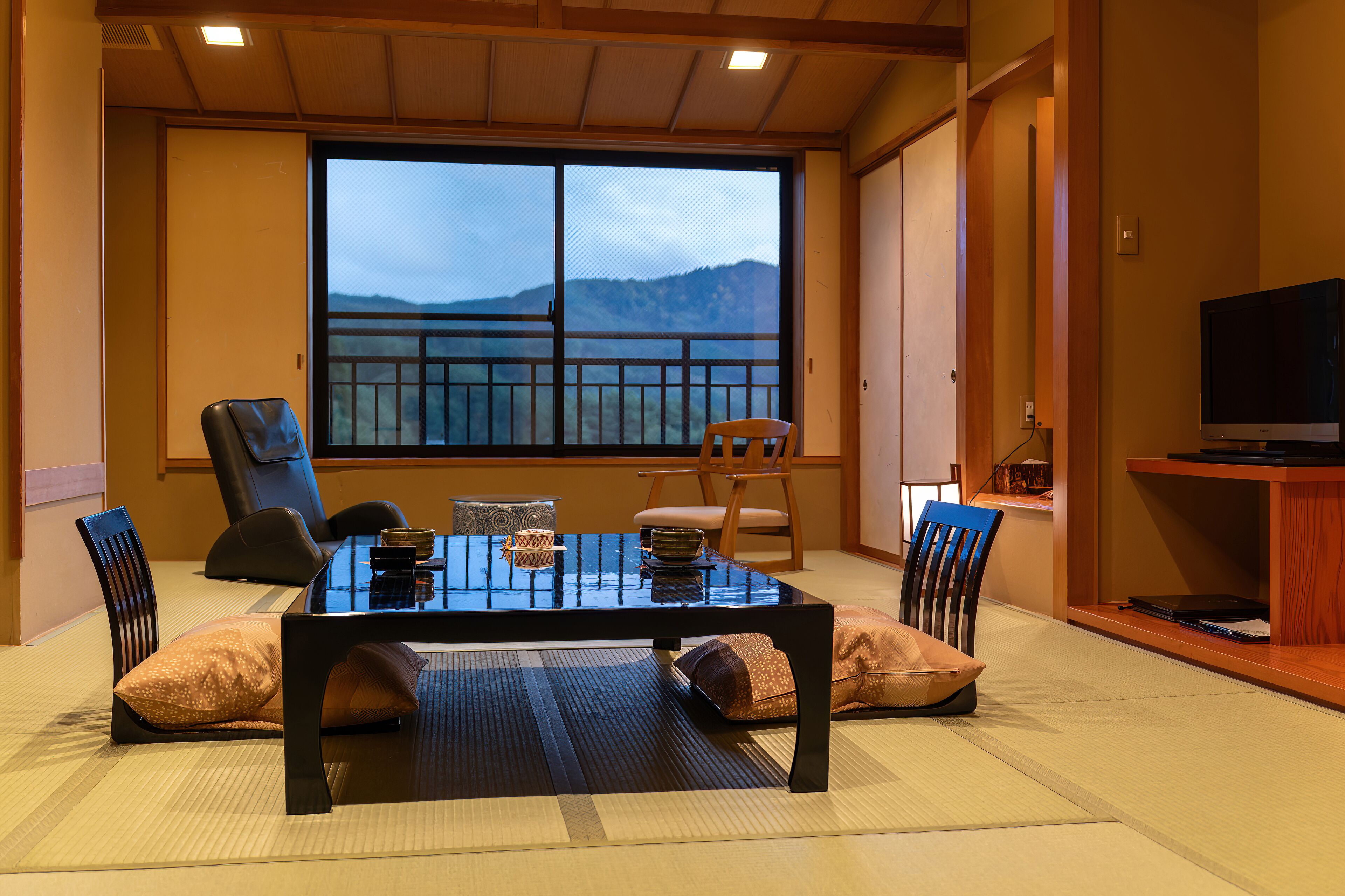 Ein traditioneller japanischer Raum mit Tatami-Boden, einem niedrigen Tisch mit Kissen und Blick auf Berge durch ein großes Fenster.