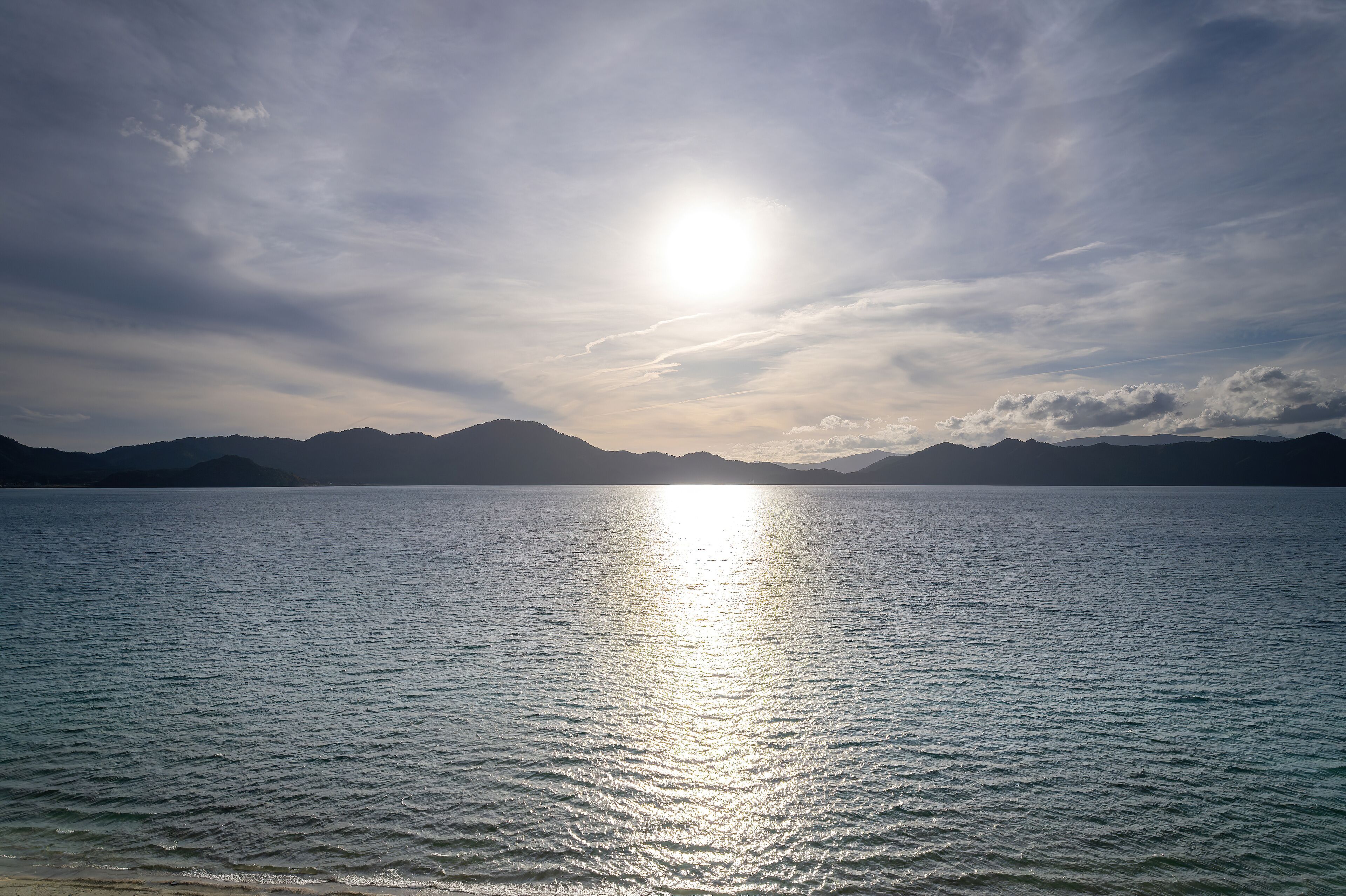 A calm lake with mountains in the background and the sun reflecting on the water.
