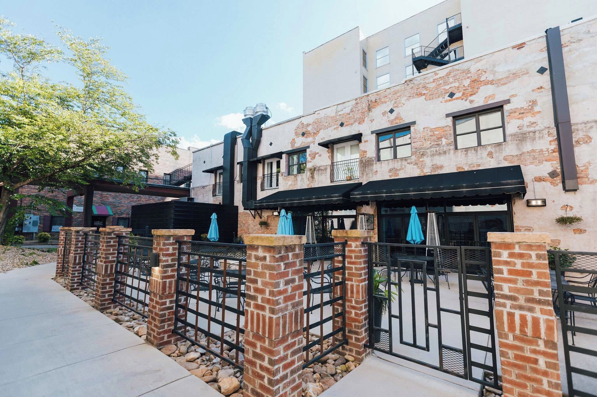 A row of brick buildings with outdoor seating areas, a gated entrance, and a clear sky.