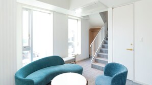 Una sala moderna con un sofá curvo color aguamarina, una mesa redonda blanca y un sillón a juego. Una escalera con barandilla de madera conduce a un piso superior.