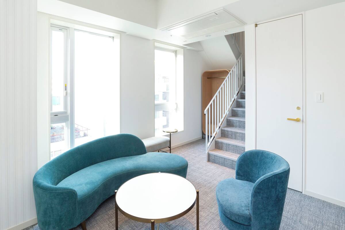 Una sala moderna con un sofá curvo color aguamarina, una mesa redonda blanca y un sillón a juego. Una escalera con barandilla de madera conduce a un piso superior.