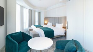 Habitación de hotel con una cama grande, dos sillones, una mesita y un amplio ventanal con cortinas.