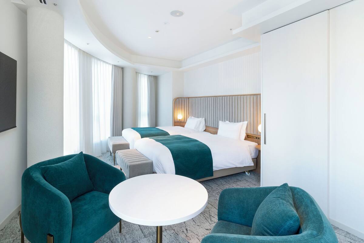 Habitación de hotel con una cama grande, dos sillones, una mesita y un amplio ventanal con cortinas.