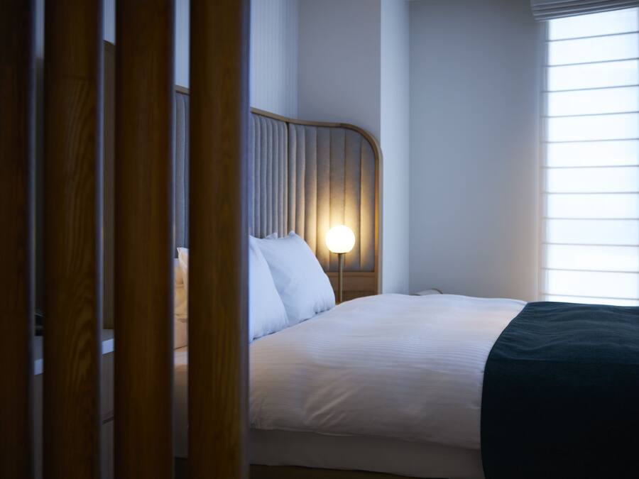 Ein ordentlich bezogenes Bett mit einem hölzernen Kopfteil, einer Nachttischlampe und einem Fenster mit Jalousien.