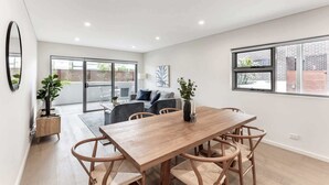 A modern dining area with a wooden table and woven chairs, a grey sofa, and a large window with a view of a brick building.