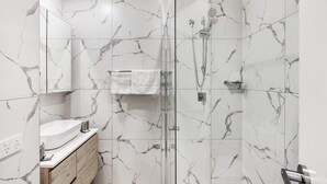 A modern bathroom with a glass shower enclosure, a wooden vanity, and marble-patterned walls. - Pristine 3-Bed Kiama Unit with Courtyard (Kiama)