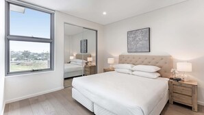 A modern bedroom with a large bed, bedside tables, a mirror, and a view through a window. - 3-Bed Apartment in Beautiful Kiama with Study Nook (Kiama)