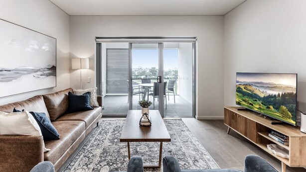 A modern living room with a sofa, coffee table, and TV. There's a balcony with a view of the city. - 3-Bed Apartment in Beautiful Kiama with Study Nook (Kiama)
