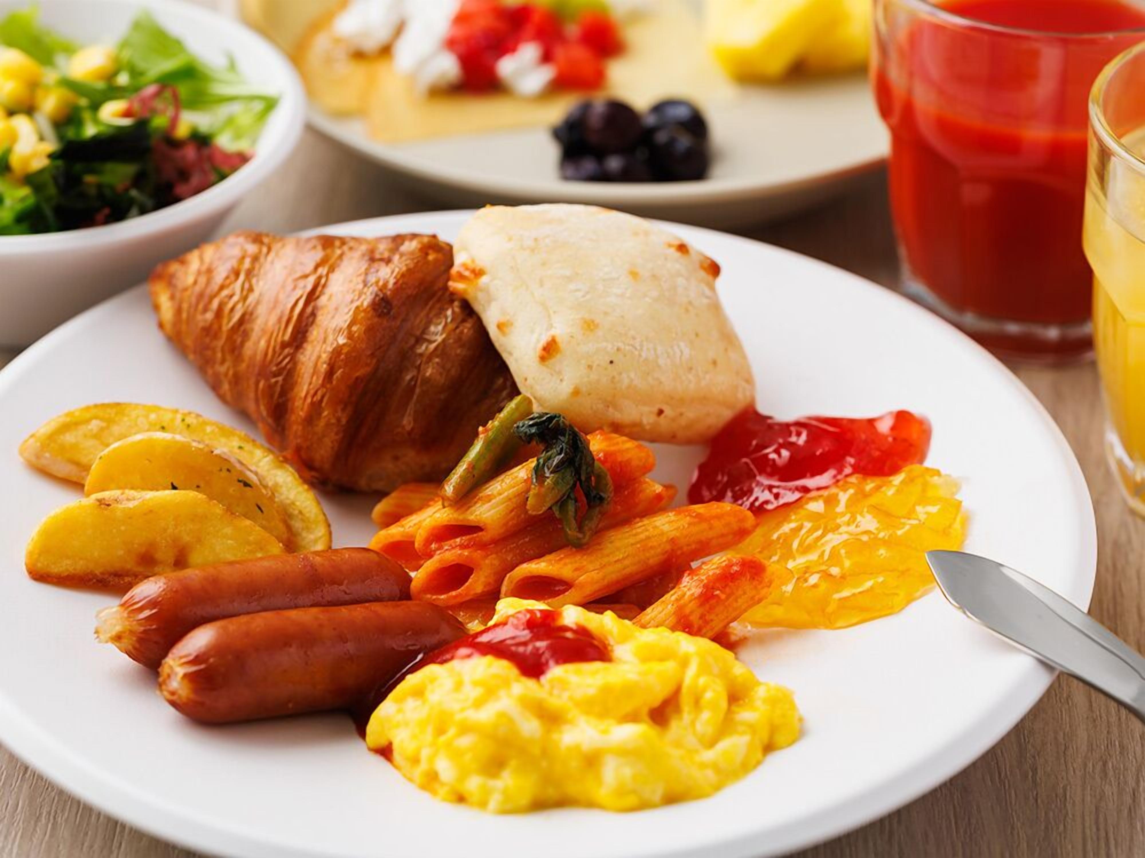 Une assiette avec des croissants, des saucisses, des pâtes, des œufs brouillés et un verre de jus de fruits.
