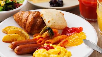 Une assiette avec des croissants, des saucisses, des pâtes, des œufs brouillés et un verre de jus de fruits.