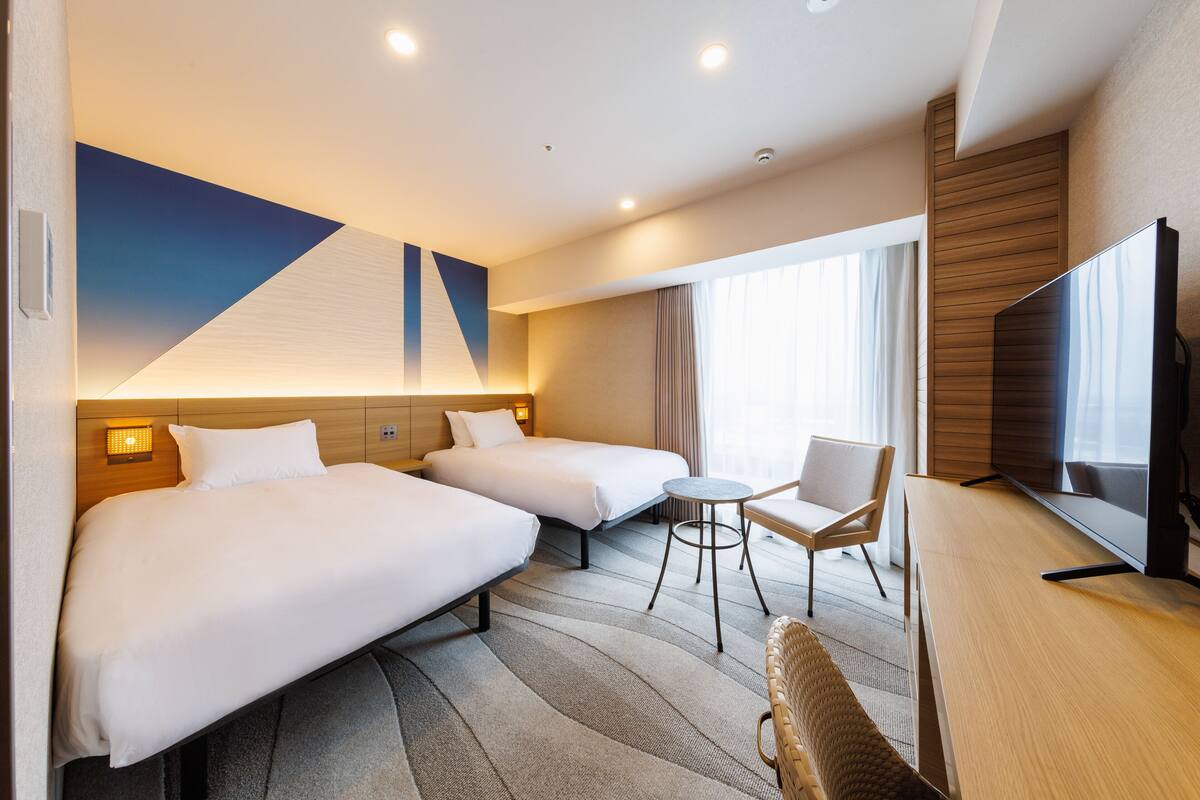 A hotel room with two beds, a small round table, a chair, a flat-screen TV, and a large window with curtains.