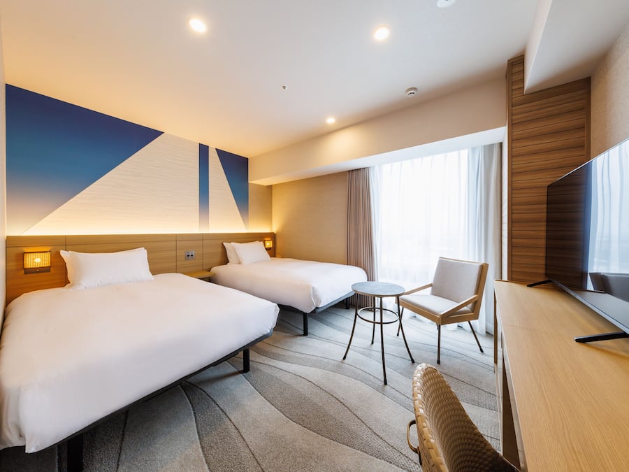 A hotel room with two beds, a small round table, a chair, a flat-screen TV, and a large window with curtains.