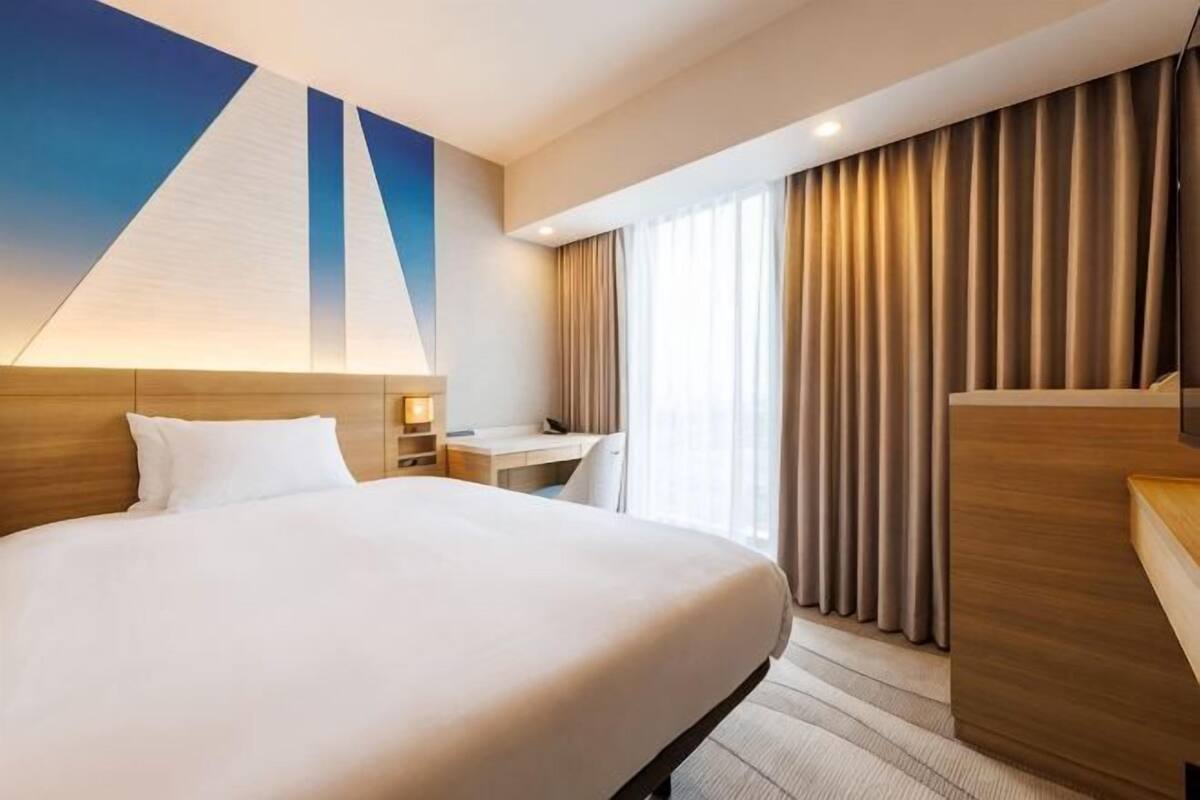 A hotel room with a large bed, a desk, and a chair, a window with curtains, and a wall with a geometric design.