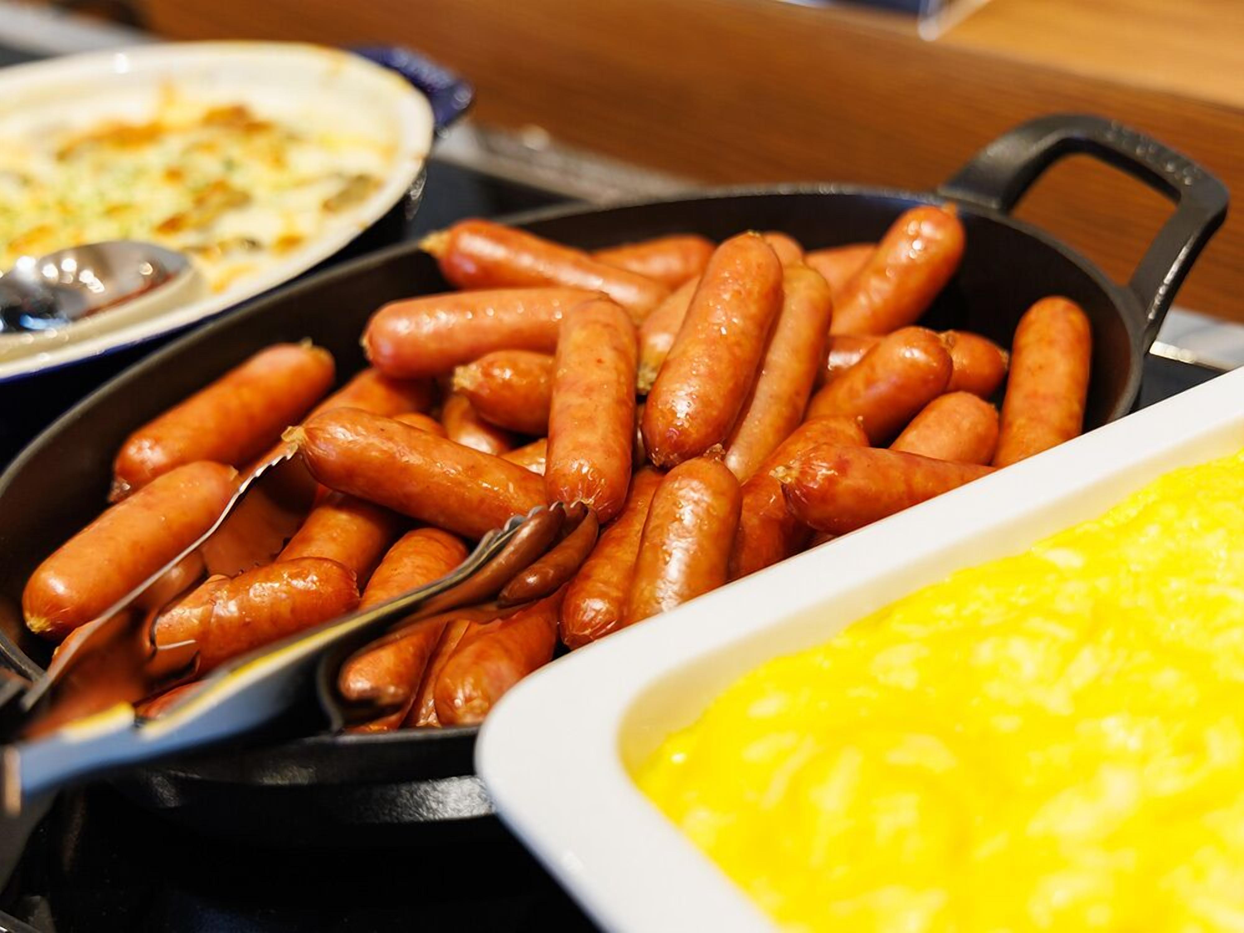 A skillet filled with sausages, a white dish with yellow substance, and a plate with food in the background.