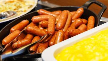 A skillet filled with sausages, a white dish with yellow substance, and a plate with food in the background.