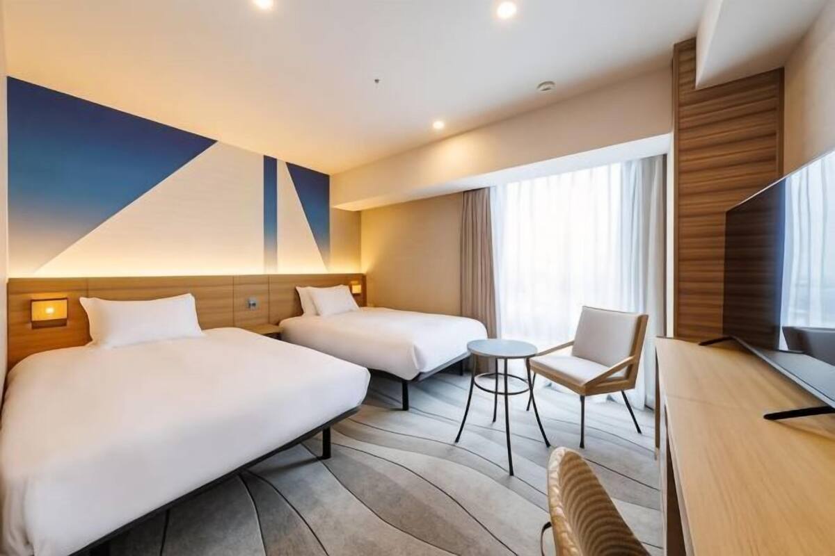 A hotel room with two beds, a small round table, a chair, a TV, and a large abstract painting on the wall.