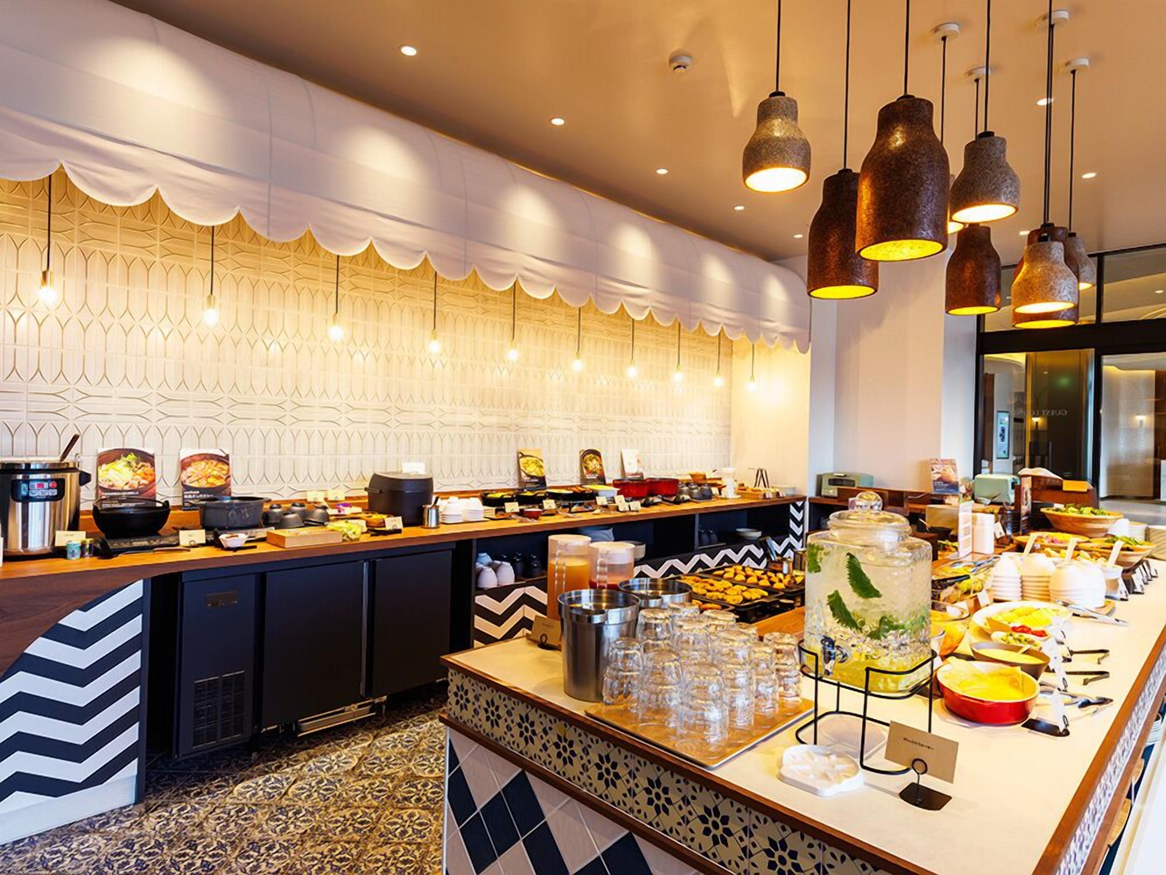 A buffet setup with various food items and beverages, a patterned wall, and hanging pendant lights.