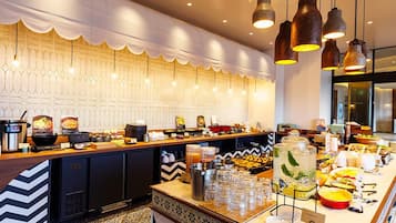 A buffet setup with various food items and beverages, a patterned wall, and hanging pendant lights.