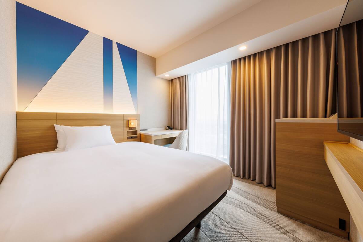 A hotel room with a large bed, a desk, a chair, a TV, and a window with curtains.