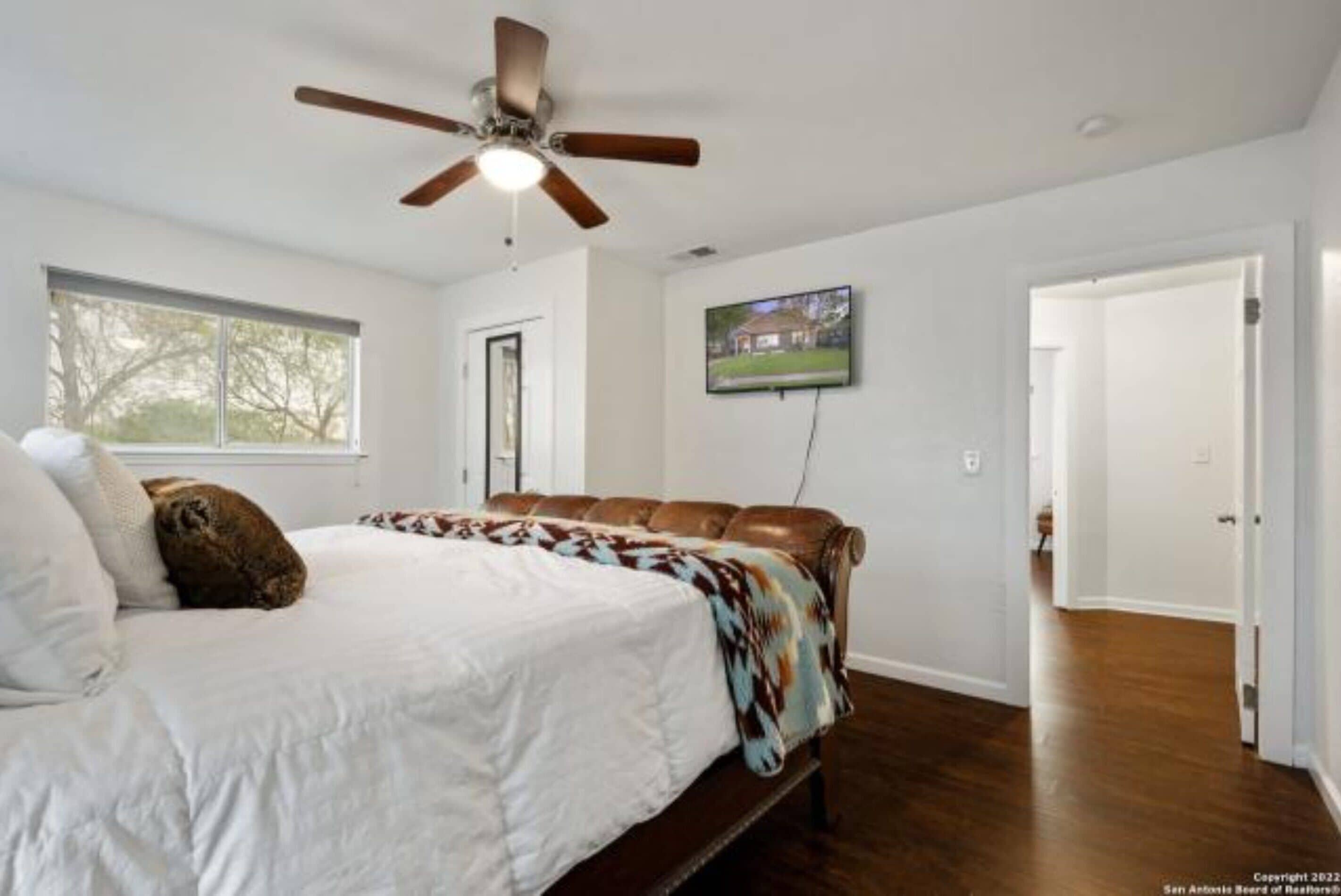 A bedroom with a bed, a ceiling fan, a television on the wall, and a window.