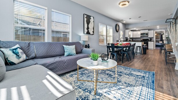 Un salon moderne avec un canapé gris, une table basse ronde et un tapis à motifs bleus. On y trouve également un coin repas avec une table et des chaises, et une cuisine à l’arrière-plan.