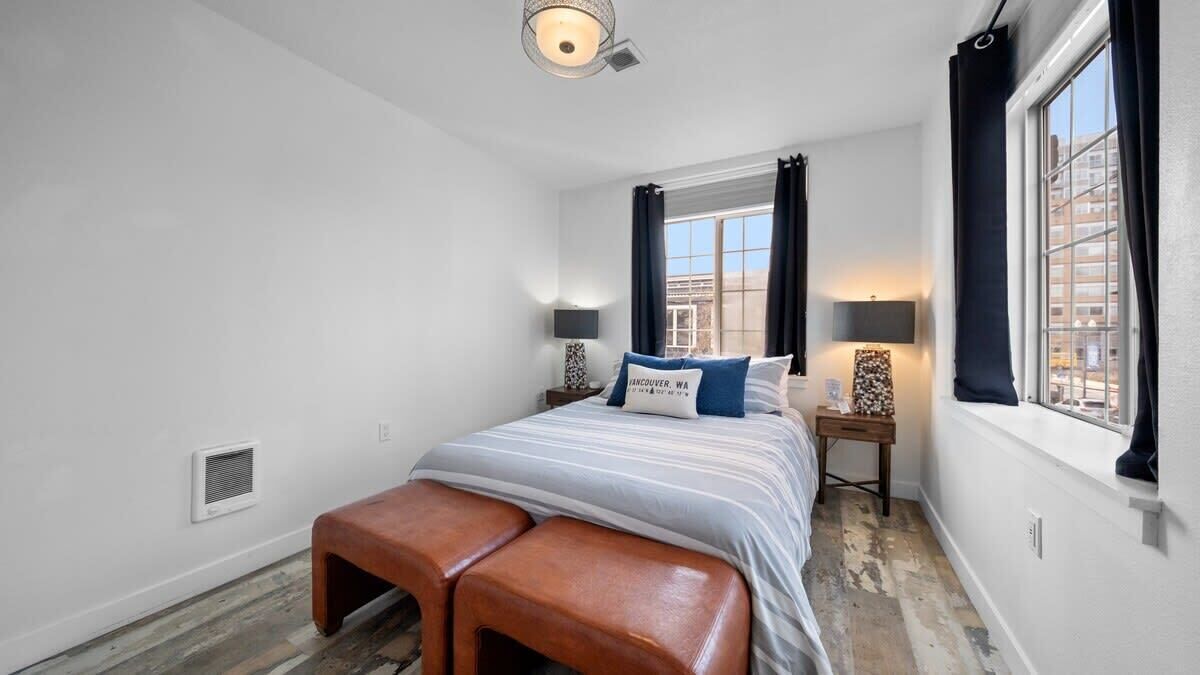 A bedroom with a bed, two bedside tables, a bench, a window with curtains, and a wall-mounted light fixture.