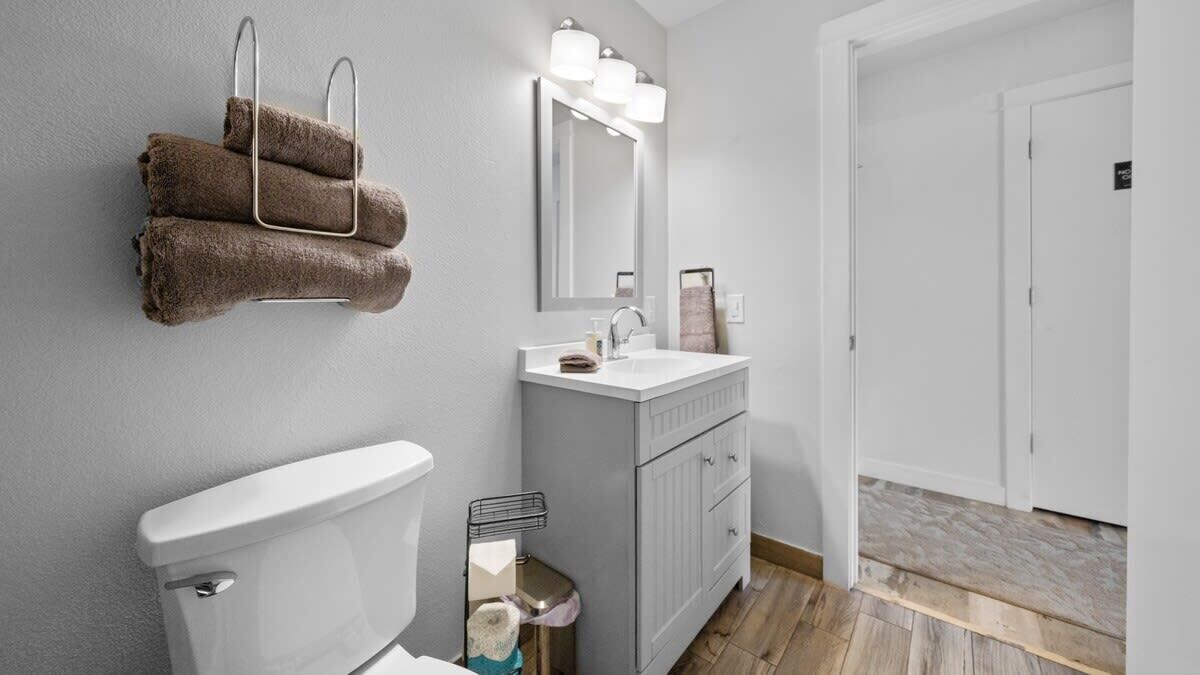 Et moderne badeværelse med toilet, vask og spejl. Der er håndklæder på en bøjle, en skraldespand og et bruseområde.