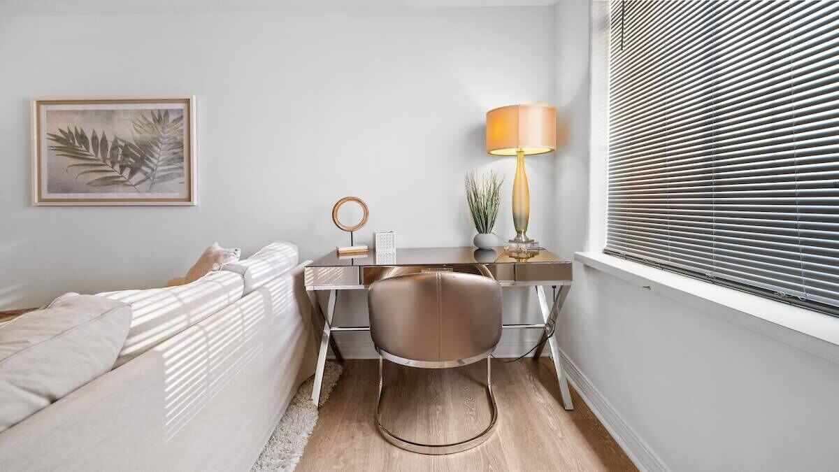 A bedroom with a bed, a desk, a chair, a lamp, a window with blinds, and a framed picture on the wall.