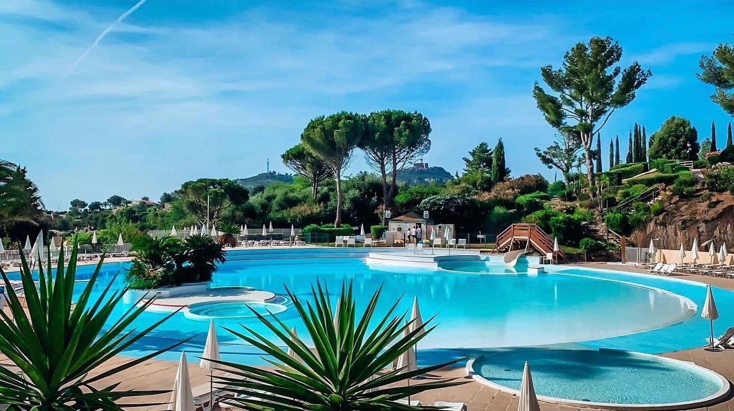 A large outdoor swimming pool with a winding design, surrounded by lounge chairs and umbrellas, set against a backdrop of trees and hills.