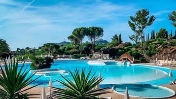 Une grande piscine extérieure à la forme sinueuse, entourée de chaises longues et de parasols, avec en arrière-plan un paysage de arbres et de collines.