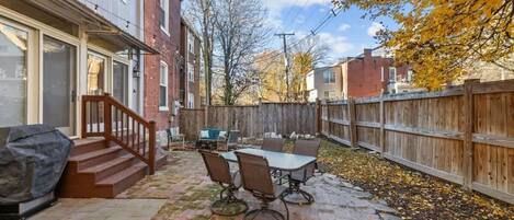 A backyard patio with a table and chairs, a covered grill, and a wooden fence.