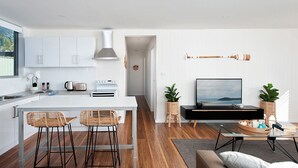 A modern kitchen with white cabinets, a central island with bar stools, a built-in oven, and a large window. - Aloha Beach House – downstairs suite, close to beach (Fingal Bay)