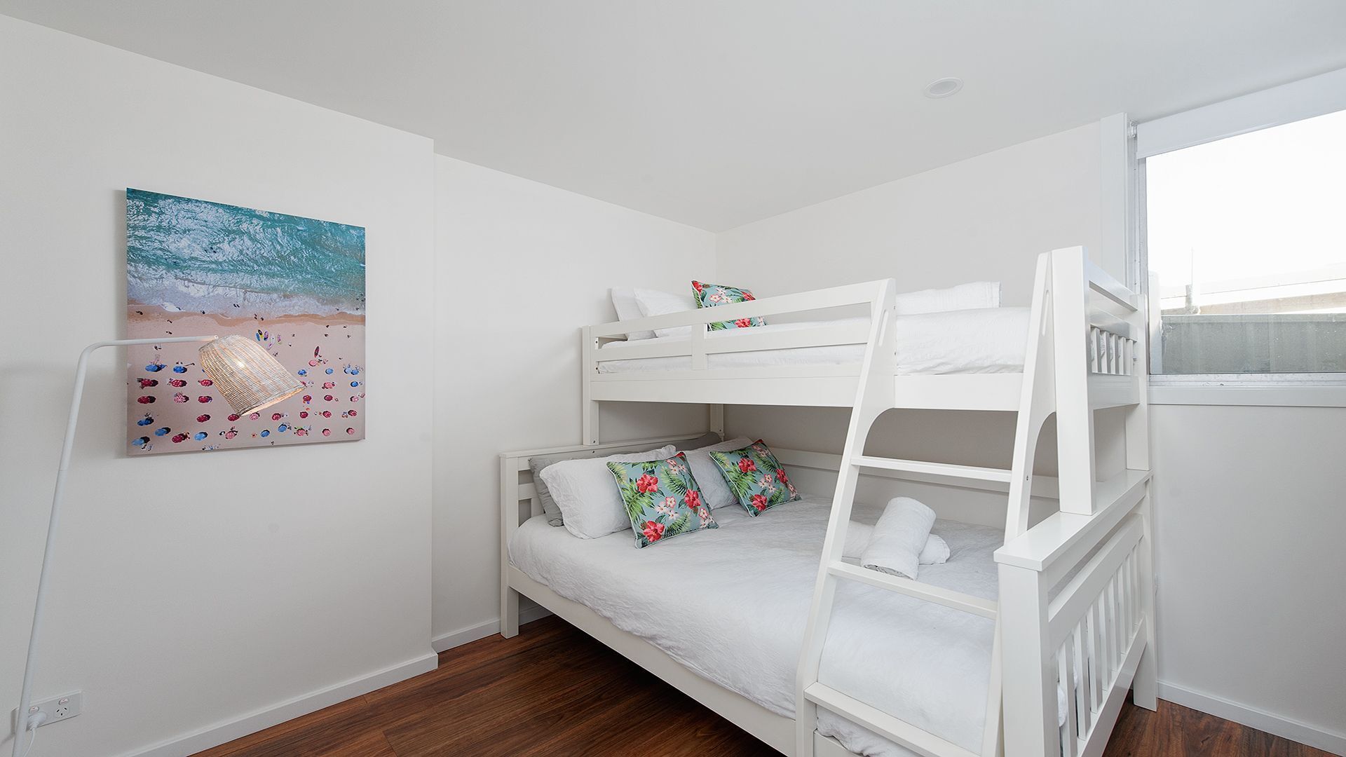 Een slaapkamer met stapelbedden, een schilderij met strandthema aan de muur, witte beddengoed en een raam met uitzicht.