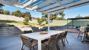 An outdoor patio with a table and chairs, a grill, and a pergola with blue glass panels. - Aloha Beach House – downstairs suite, close to beach (Fingal Bay)