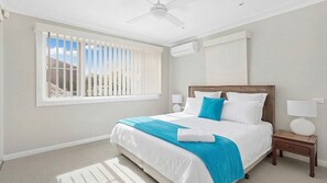 A bedroom with a bed, a window with blinds, a ceiling fan, and a nightstand with a lamp. - Bay Escape — ocean views, close to cafes (Nelson Bay)