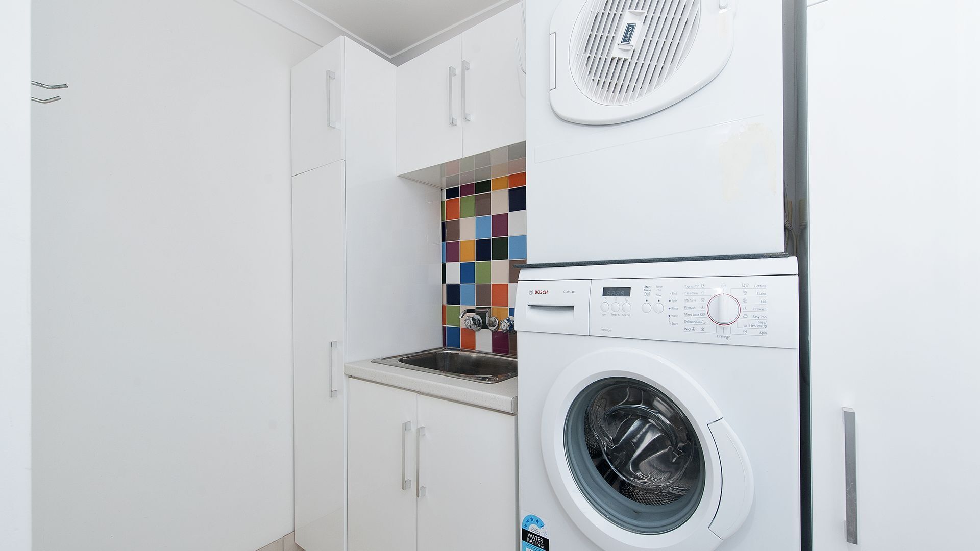 A compact laundry room with a Bosch washing machine, a sink, and a colorful tile backsplash.