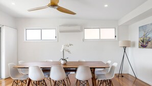 Une salle à manger moderne avec une table en bois, des chaises blanches, un ventilateur de plafond et un tableau accroché au mur.