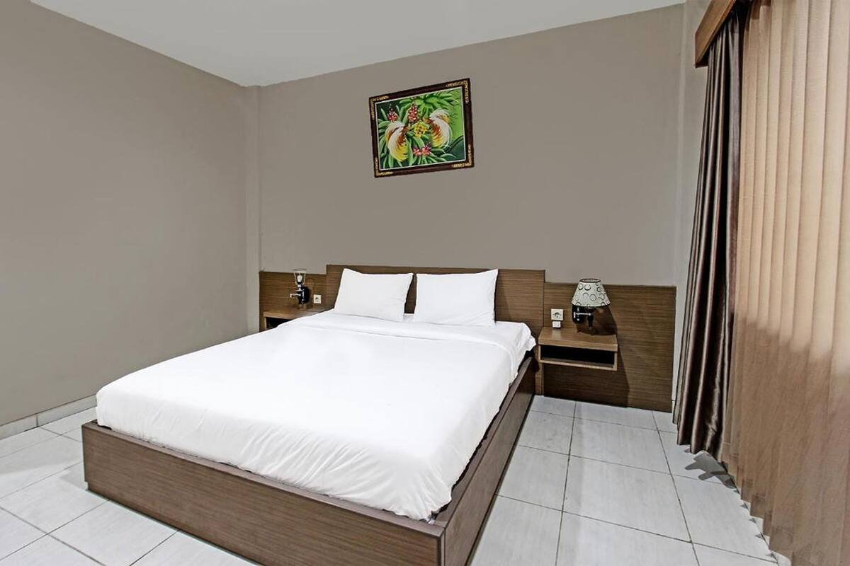 A hotel room with a large bed, wooden headboard, white bedding, a nightstand with a lamp, and a wall-mounted picture.