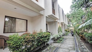 A narrow pathway flanked by white buildings with balconies, surrounded by lush greenery and a parked motorcycle. - Kimara Homestay (Senggigi)