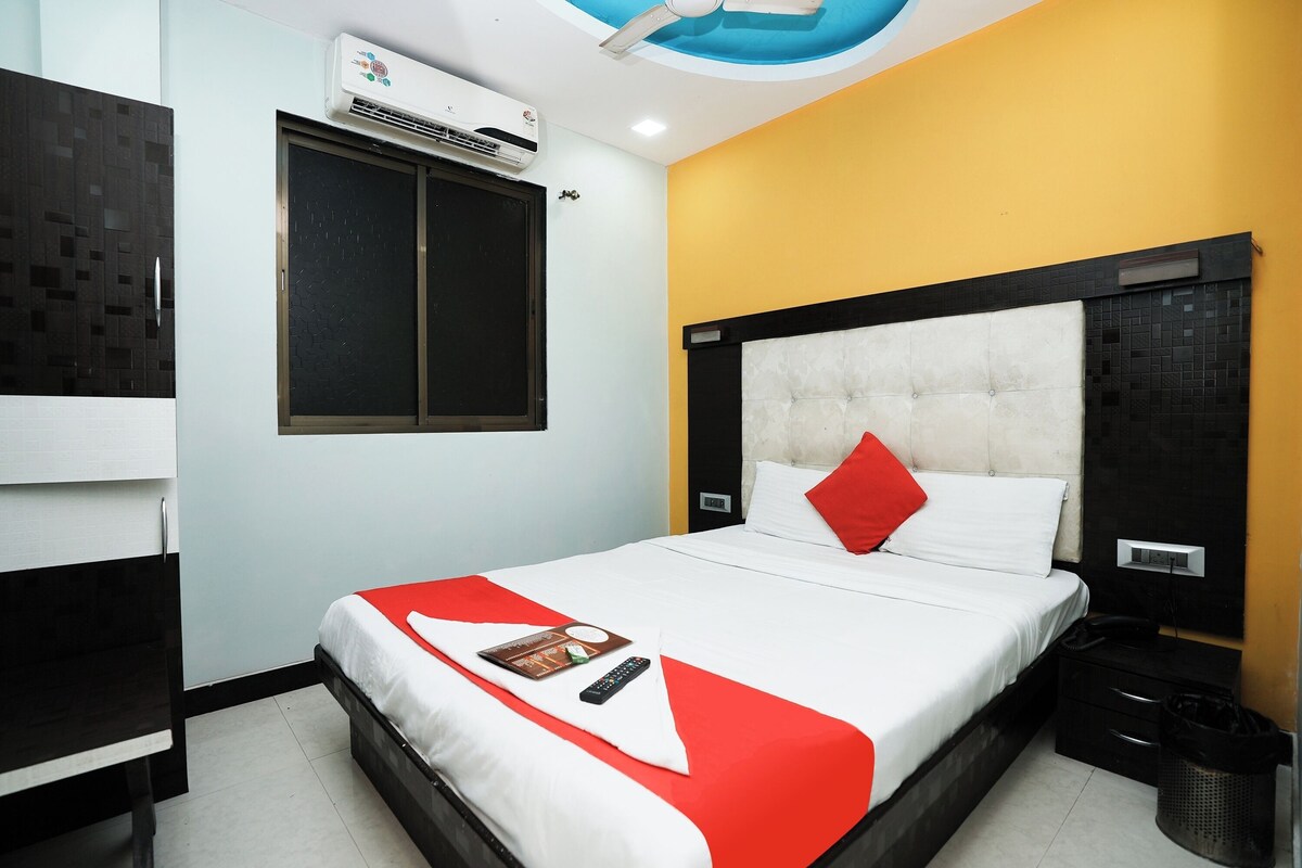 A hotel room with a bed, a window, an air conditioning unit, a bedside table, and a trash bin.