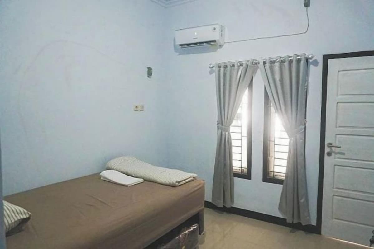 A small, single-bed room with a window, an air conditioning unit, and a door.