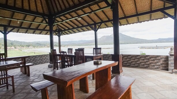 An outdoor seating area with wooden tables and benches, a tiled floor, and a view of a body of water and mountains in the background. - Baruna Lakeside View (Kintamani)