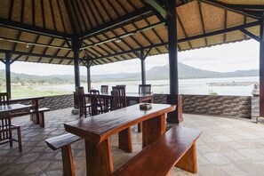 An outdoor seating area with wooden tables and benches, a tiled floor, and a view of a body of water and mountains in the background. - Baruna Lakeside View (Kintamani)